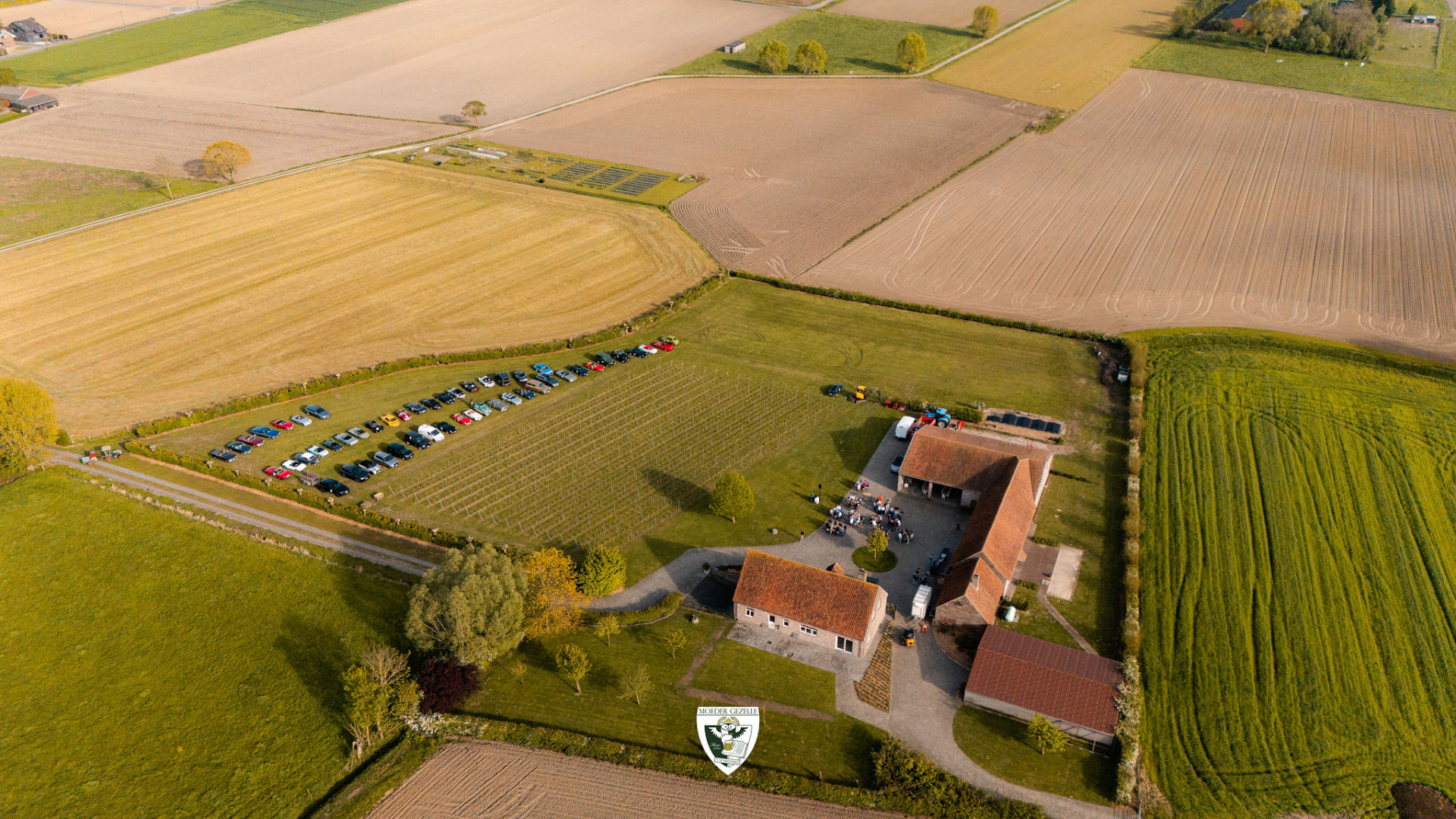 Luchtbeeld van de hoeve met een lange rij geparkeerde oldtimers langs de velden