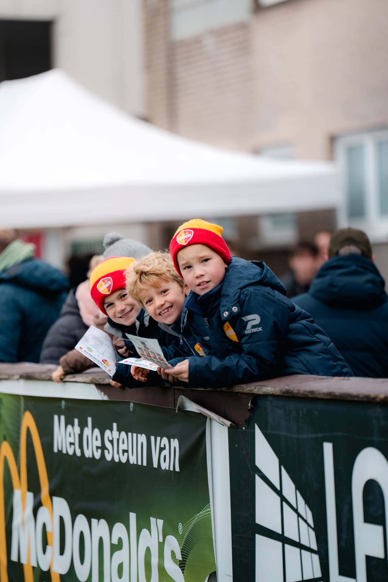 Twee jonge supporters met rood-gele muts leunen lachend over de omheining