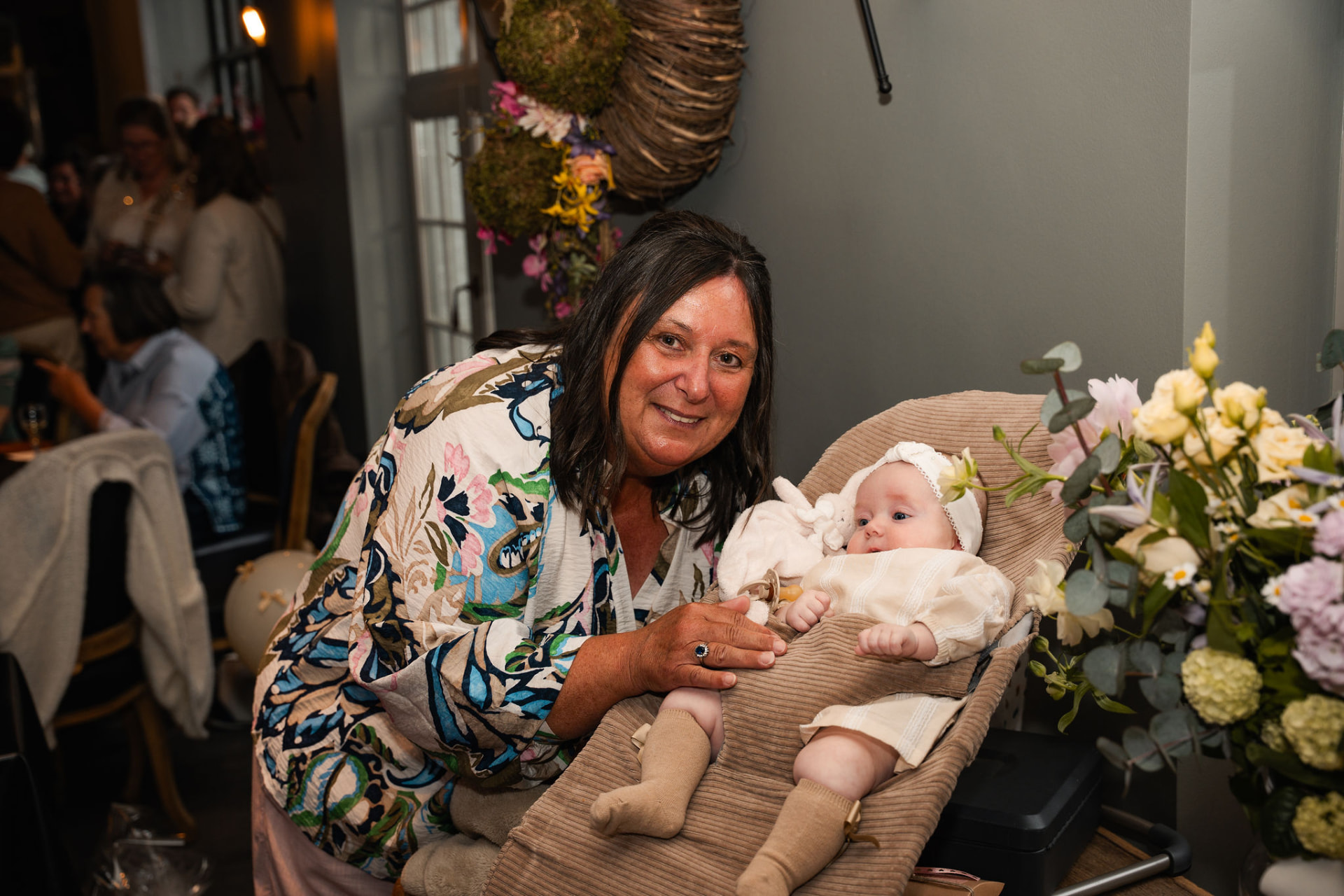 Oma poseert lachend naast Manon in haar wipstoeltje bij de bloemen