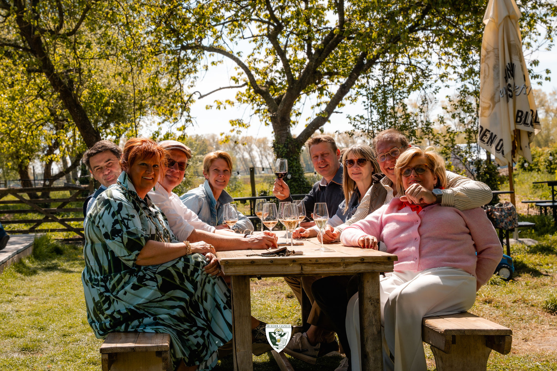 Groep gasten aan een picknicktafel onder de bomen met glazen wijn
