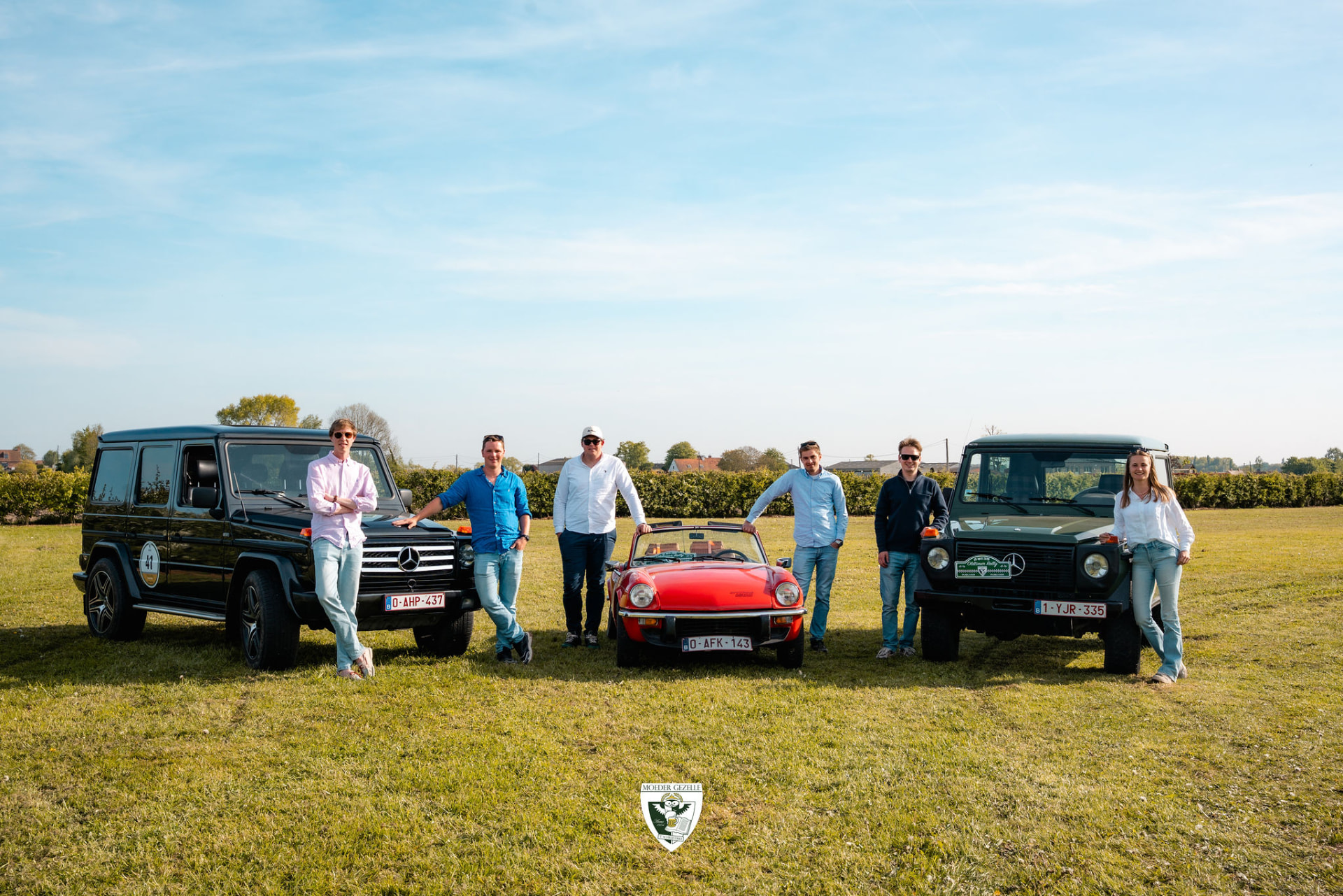 Groep deelnemers poseert bij twee terreinwagens en een rode Triumph Spitfire